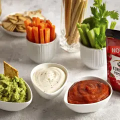 Trzy miseczki z dipami: guacamole, śmietanowym i pomidorowym, obok warzyw i krakersów.