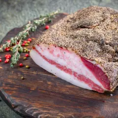 Guanciale co to? Włoski, peklowany policzek wieprzowy, pokryty pieprzem, leży na drewnianej desce z ziołami i pieprzem.
