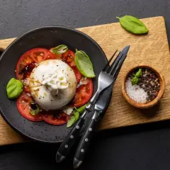 Pyszna burrata z pomidorami, polana oliwą, ozdobiona bazylią. Obok miseczka z solą i pieprzem.
