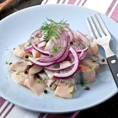 Kawałki śledzia z cebulą i koperkiem na jasnoniebieskim talerzu, obok widelec.