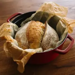 Złocisty bochenek chleba na zakwasie w czerwonym naczyniu żeliwnym, wyłożonym papierem do pieczenia.