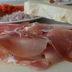 Cienko pokrojona szynka crudo, kawałek parmezanu i talerz z dodatkami.