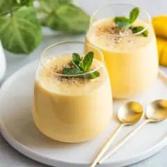 Dwa szklanki gęstego, żółtego smoothie mango z miętą i posypką, obok kawałka mango i łyżeczek.
