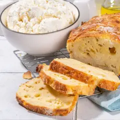 Chleb z twarogu, świeżo upieczony, z chrupiącą skórką i puszystym wnętrzem. Obok miska pełna białego twarogu, składnika tego pysznego wypieku.