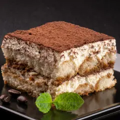 Kawałek tiramisu posypany kakao, z listkami mięty i ziarnami kawy na czarnym talerzu.