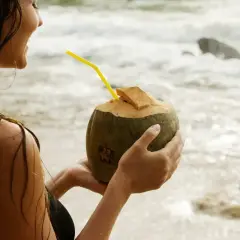 Uśmiechnięta kobieta na plaży trzyma kokosa z żółtą słomką. Woda kokosowa właściwości nawadniające i odżywcze.