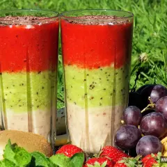 Dwa warstwowe koktajle owocowe w szklankach, otoczone owocami: kiwi, truskawki, winogrona i banan.