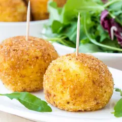 Dwa złociste arancini z wykałaczkami na białym talerzu, obok sałatka.