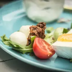 Keto śniadanie: ser brie z dżemem, kulki mozzarelli, pomidor i rukola na niebieskim talerzu.