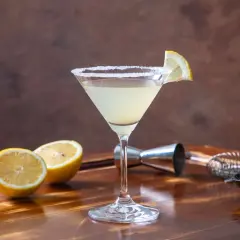 Elegancki martini bianco drink z solnym rantem, ozdobiony plasterkiem cytryny. Obok leżą połówki cytryn i miseczka oliwek.