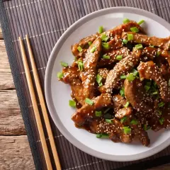 Pokrojona wołowina w sosie teriyaki, posypana sezamem i szczypiorkiem, na talerzu z pałeczkami.