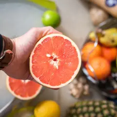 Dłoń trzyma przekrojonego grejpfruta, obok inne owoce. To zdrowa przekąska, niskokaloryczny grejpfrut.