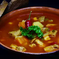 Gęsty bogracz wieprzowy z kawałkami mięsa, ziemniaków i papryki, ozdobiony papryczką chili i natką pietruszki. Obok kromki chleba.