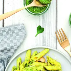 Makaron penne z zielonym pesto, podany na talerzu, obok słoików z pesto i złotego widelca.