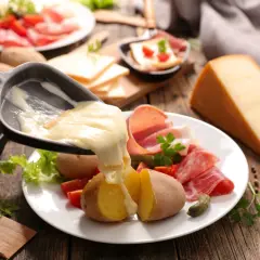 Roztopiony ser raclette spływa na ziemniaki i wędliny. Obok kawałek sera i talerz z dodatkami.