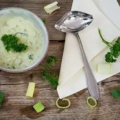 Kremowy sos porowy z natką pietruszki, podany w misce, obok łyżki i kawałków pora na drewnianym stole.