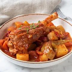 Pieczona goleń jagnięca w sosie z warzywami i ziemniakami w misce.