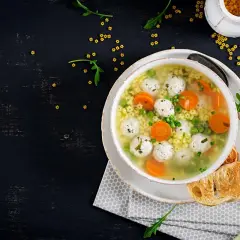 Miseczka zupy z klopsikami, makaronem, marchewką i groszkiem na czarnym tle.