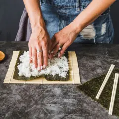 Dłonie rozkładają ryż na nori na macie bambusowej do sushi.