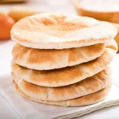 Stos świeżo upieczonego chleba arabskiego na białej serwetce, obok jajka i wałka do ciasta.