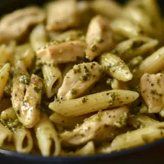Makaron penne z kawałkami kurczaka w sosie pesto, podany w ciemnej misce.