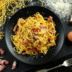 Pyszne spaghetti carbonara na talerzu, obok surowy boczek, jajko i ser. Zastanawiasz się, ile kalorii ma spaghetti carbonara?