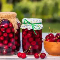 Dwa słoiki kompotu z czereśni i miseczka świeżych czereśni na stole.
