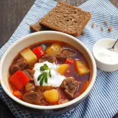 Gęsty, aromatyczny **бограч** z kawałkami wołowiny, ziemniaków i papryki, udekorowany śmietaną i natką pietruszki. Obok kromka chleba i miseczka śmietany.