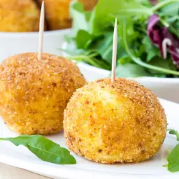 Dwa złociste arancini z wykałaczkami na białym talerzu, obok sałatka.