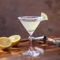 Elegancki martini bianco drink z solnym rantem, ozdobiony plasterkiem cytryny. Obok leżą połówki cytryn i miseczka oliwek.