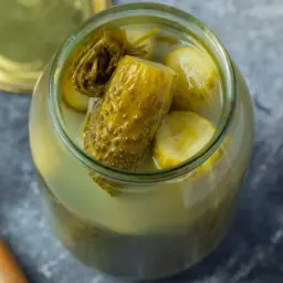 Słoik pełen kiszonych ogórków w aromatycznej wodzie z ogórków kiszonych. Złota pokrywka obok.