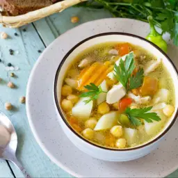 Aromatyczna zupa na rosole z kurczakiem, ciecierzycą, marchewką i ziemniakami, udekorowana natką pietruszki.