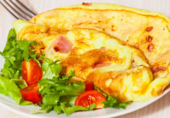 Złocisty omlet z szynką i pomidorkami koktajlowymi na talerzu z sałatą.