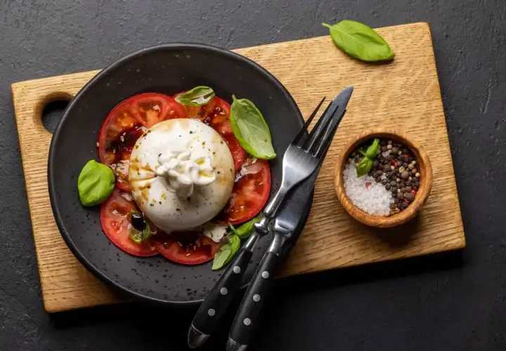 Pyszna burrata z pomidorami, polana oliwą, ozdobiona bazylią. Obok miseczka z solą i pieprzem.
