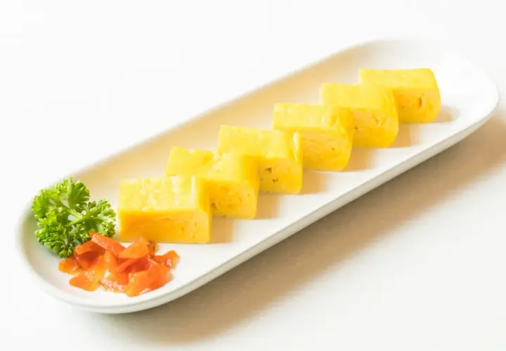 Japoński omlet tamagoyaki pokrojony w plastry, podany z natką pietruszki i marynowaną marchewką.