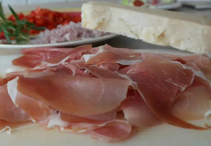 Cienko pokrojona szynka crudo, kawałek parmezanu i talerz z dodatkami.