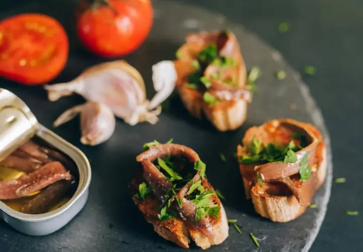 Otwarta puszka anchois, bruschetty z anchois i natką pietruszki, pomidory i czosnek na ciemnym tle.