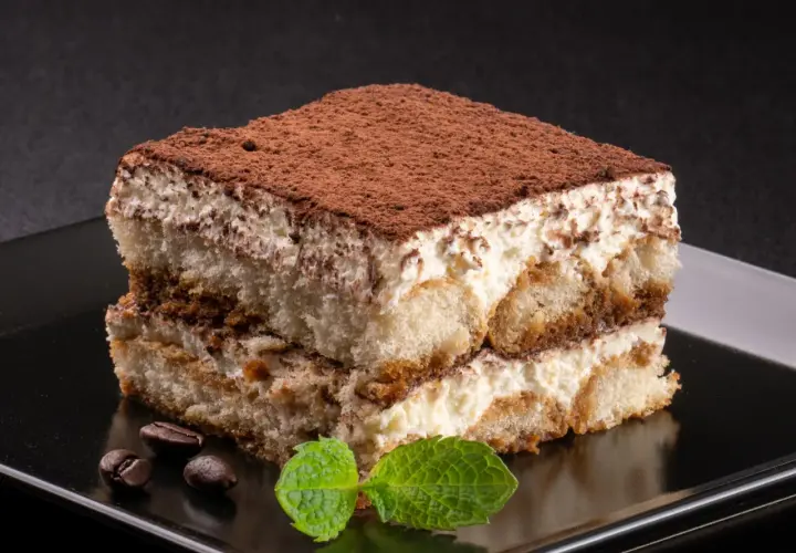 Kawałek tiramisu posypany kakao, z listkami mięty i ziarnami kawy na czarnym talerzu.