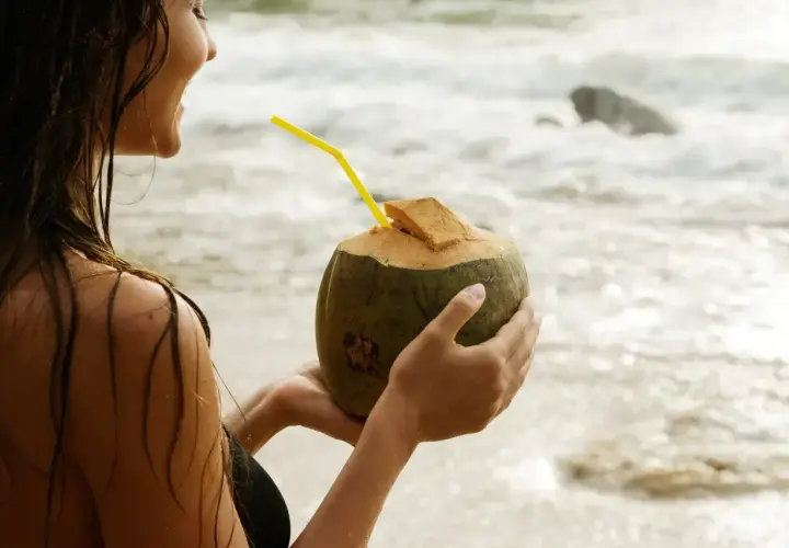 Uśmiechnięta kobieta na plaży trzyma kokosa z żółtą słomką. Woda kokosowa właściwości nawadniające i odżywcze.