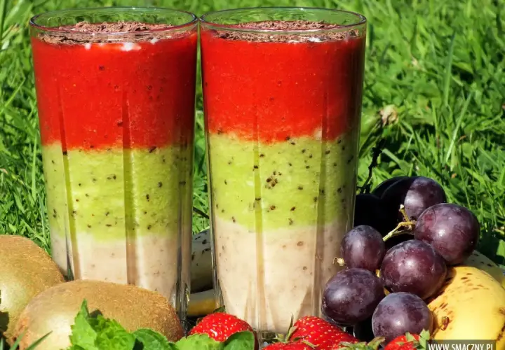 Dwa warstwowe koktajle owocowe w szklankach, otoczone owocami: kiwi, truskawki, winogrona i banan.