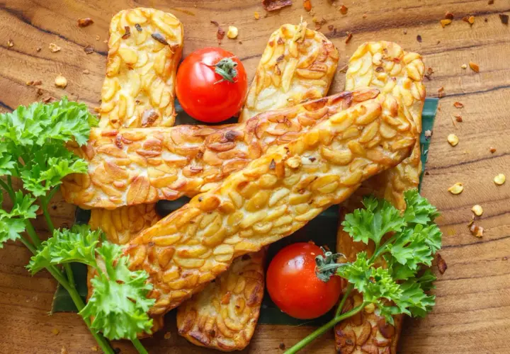 Usmażony tempeh z pomidorkami koktajlowymi i natką pietruszki na drewnianym tle.