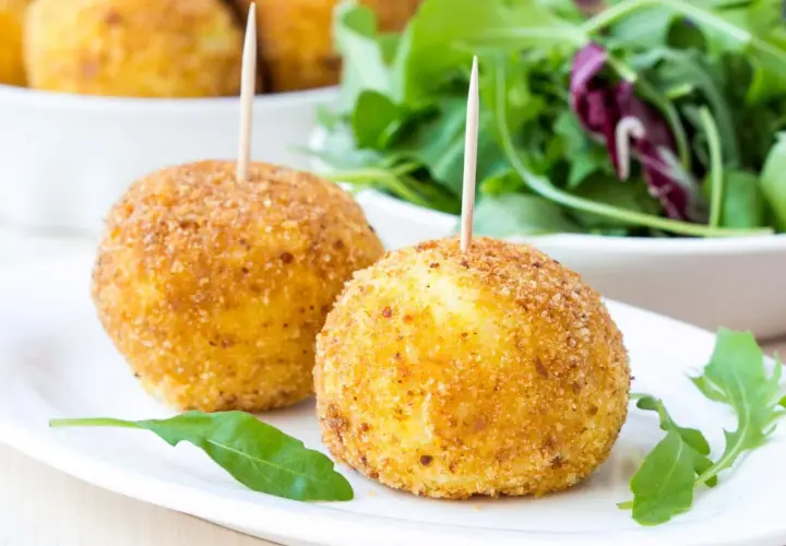 Dwa złociste arancini z wykałaczkami na białym talerzu, obok sałatka.