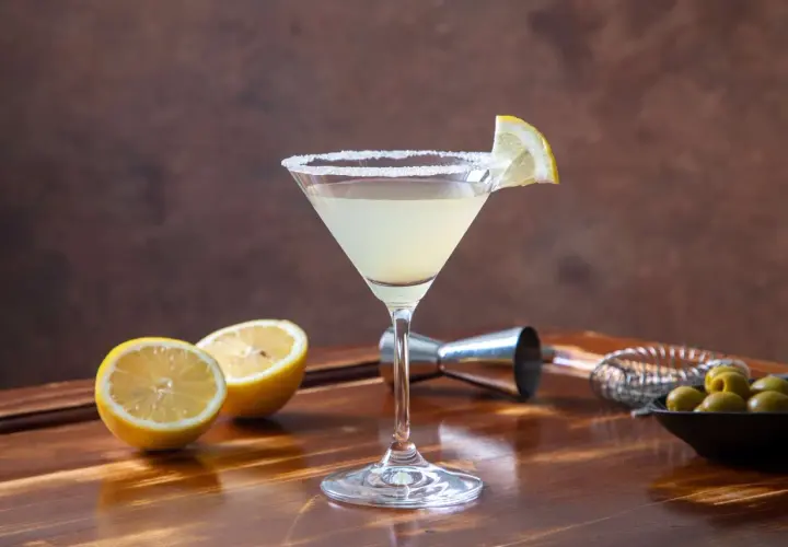 Elegancki martini bianco drink z solnym rantem, ozdobiony plasterkiem cytryny. Obok leżą połówki cytryn i miseczka oliwek.