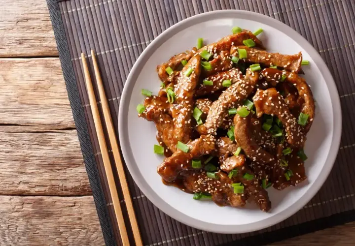 Pokrojona wołowina w sosie teriyaki, posypana sezamem i szczypiorkiem, na talerzu z pałeczkami.