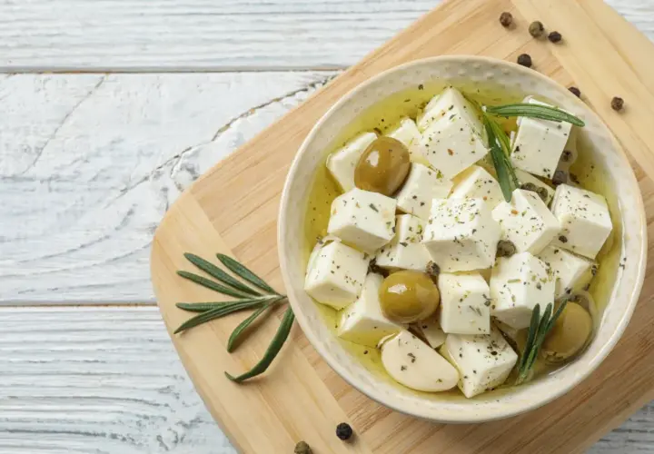 Kostki sera feta w oliwie z oliwkami, czosnkiem i rozmarynem na desce.