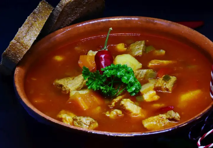 Gęsty bogracz wieprzowy z kawałkami mięsa, ziemniaków i papryki, ozdobiony papryczką chili i natką pietruszki. Obok kromki chleba.