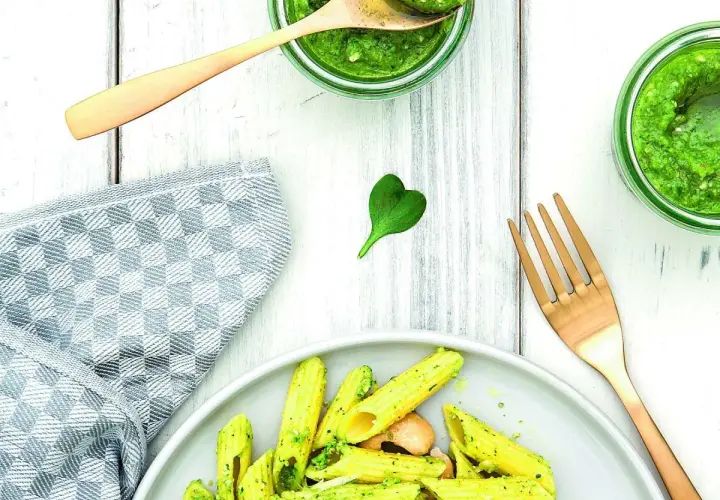 Makaron penne z zielonym pesto, podany na talerzu, obok słoików z pesto i złotego widelca.