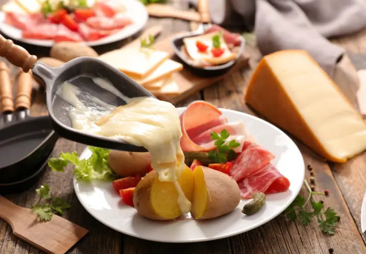Roztopiony ser raclette spływa na ziemniaki i wędliny. Obok kawałek sera i talerz z dodatkami.