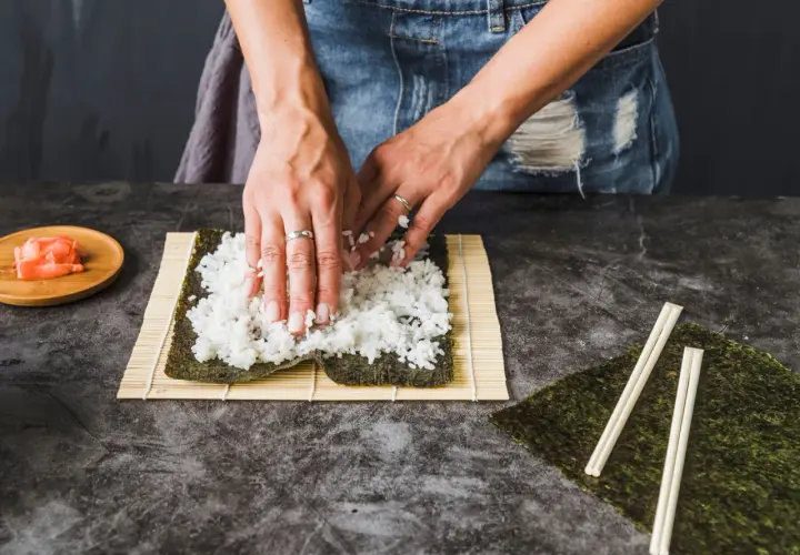 Dłonie rozkładają ryż na nori na macie bambusowej do sushi.
