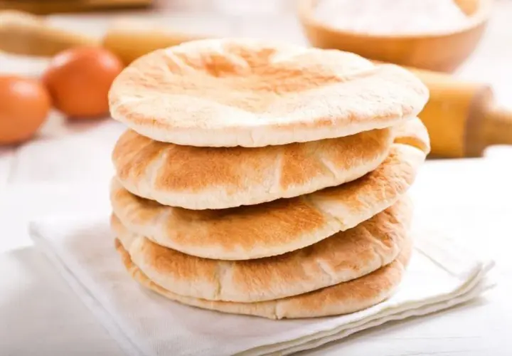 Stos świeżo upieczonego chleba arabskiego na białej serwetce, obok jajka i wałka do ciasta.